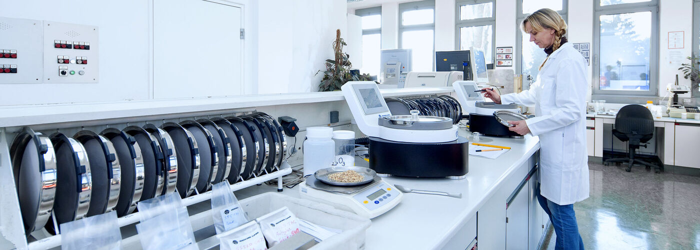  Naukowiec w fartuchu laboratoryjnym pracuje przy analizatorze z wyświetlaczem. Przed nią znajduje się waga z próbką materiału; w tle znajduje się inny sprzęt laboratoryjny.